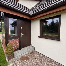 EFECT SP. Z O.O. - Brązowe okno PCV z parapetem z konglomeratu oraz ciemnobrązowe drzwi wejściowe w jasnym domu z kamiennym podłożem i małą rośliną w doniczce.