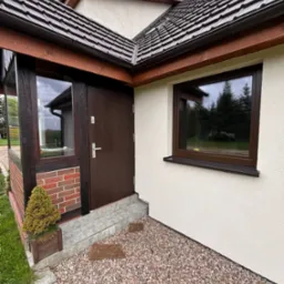 Brązowe okno PCV z parapetem z konglomeratu oraz ciemnobrązowe drzwi wejściowe w jasnym domu z kamiennym podłożem i małą rośliną w doniczce.