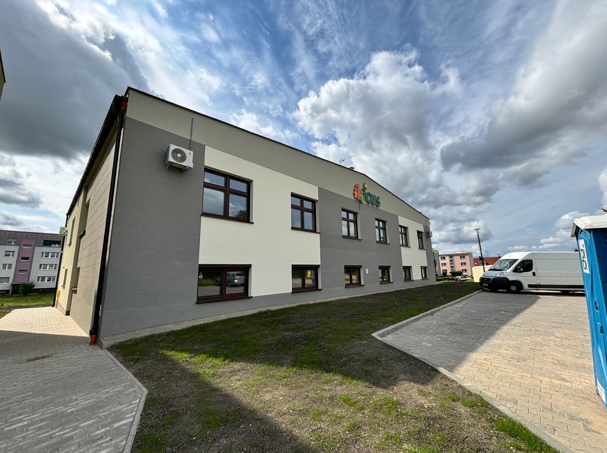 Dwukondygnacyjny budynek z szarymi i białymi ścianami, brązowymi oknami, klimatyzatorem na ścianie i logo firmy na dachu, z widocznym fragmentem parkingu i trawnika.