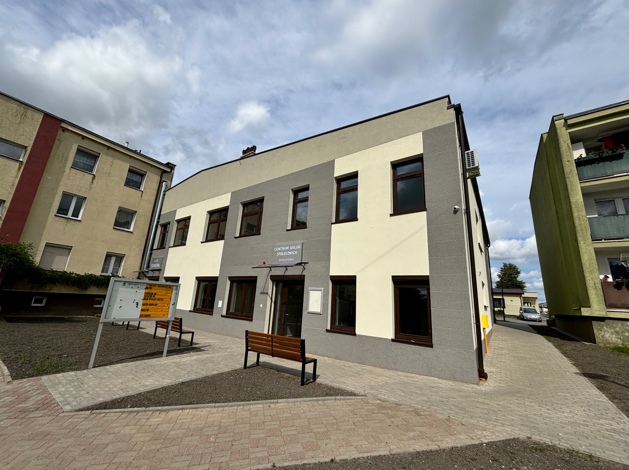 Budynek Centrum Usług Społecznych w Pelczycach z nowymi, brązowymi oknami, widok z zewnątrz, z chodnikiem i ławkami.