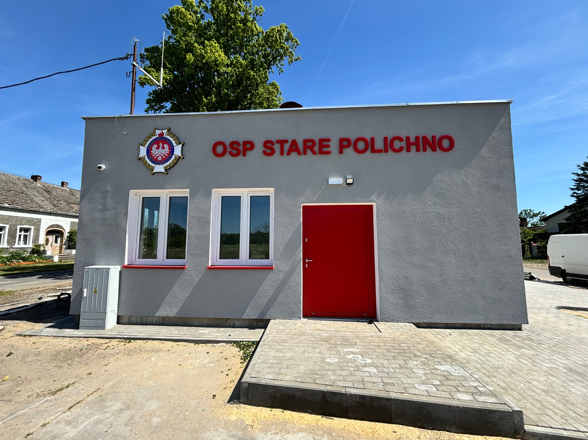 Nowo wybudowany budynek OSP w Starym Polichnie z czerwonymi drzwiami wejściowymi i białymi oknami, widok z zewnątrz w słoneczny dzień.