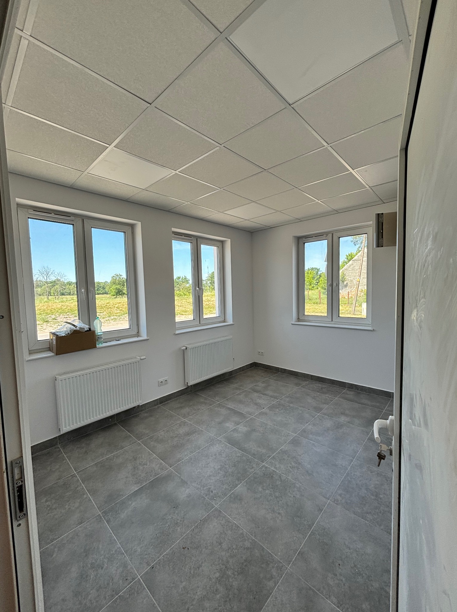Wnętrze pustego pomieszczenia z trzema białymi oknami, szarą podłogą z płytek, białymi ścianami i grzejnikami, widok na zewnątrz przez okna na zielone pole i drzewa, oraz częściowo widoczne drzwi...