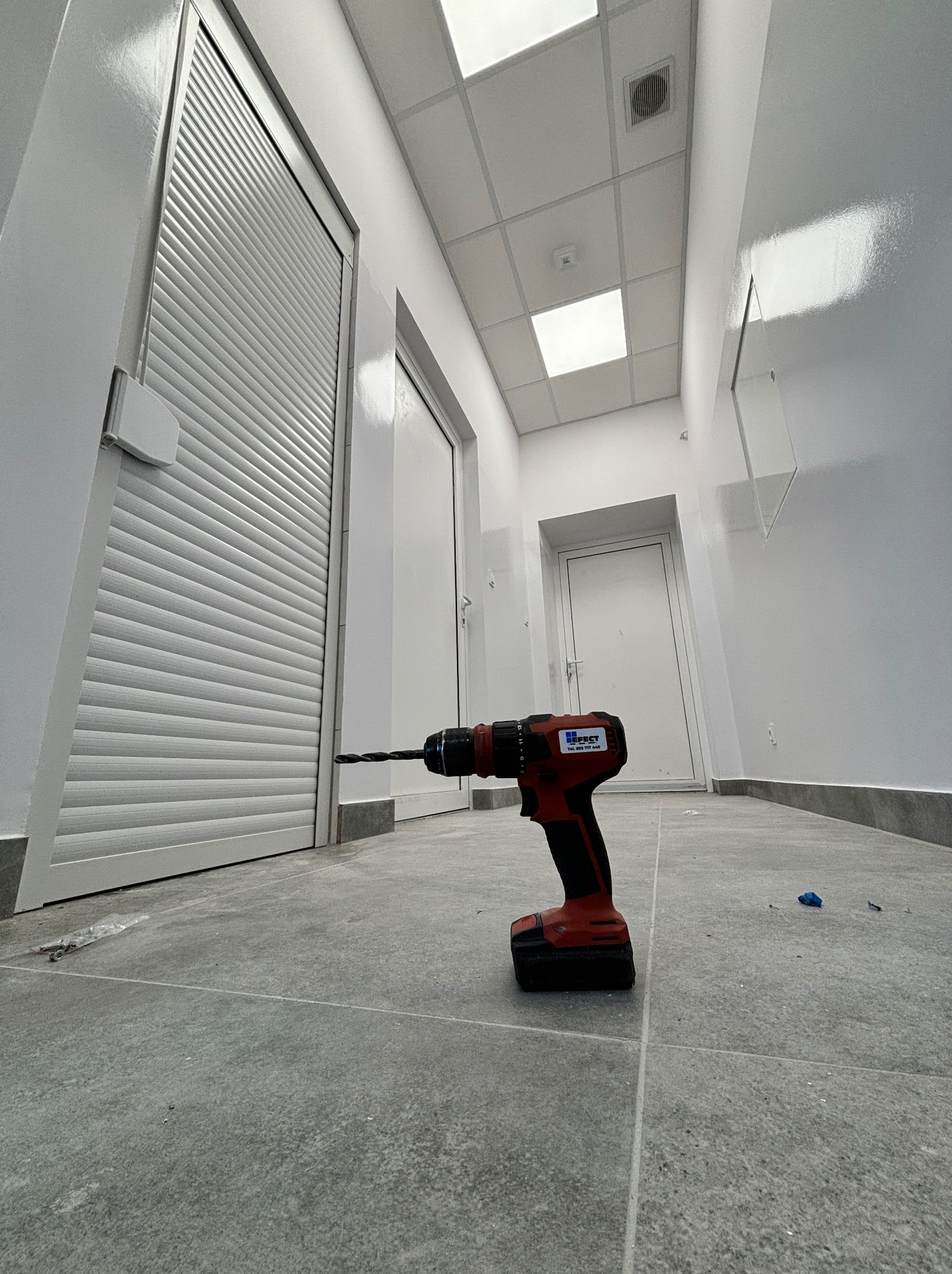 Wiertarka akumulatorowa na szarych płytkach podłogowych, na tle białych ścian i drzwi, w tym jednych z żaluzjami.