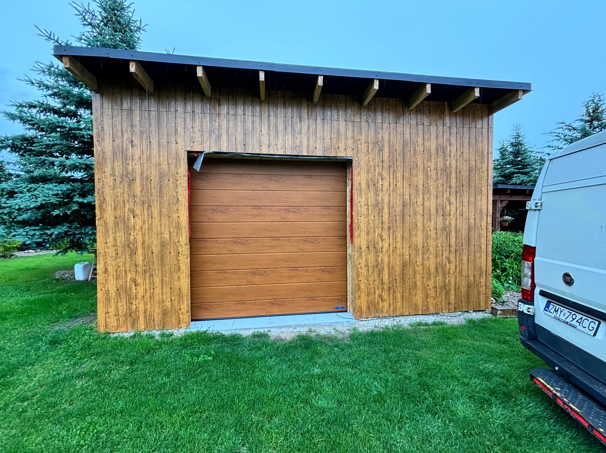 Nowo wybudowany garaż z drewnianą elewacją i segmentową bramą w kolorze drewna, częściowo widoczny samochód dostawczy na trawniku.
