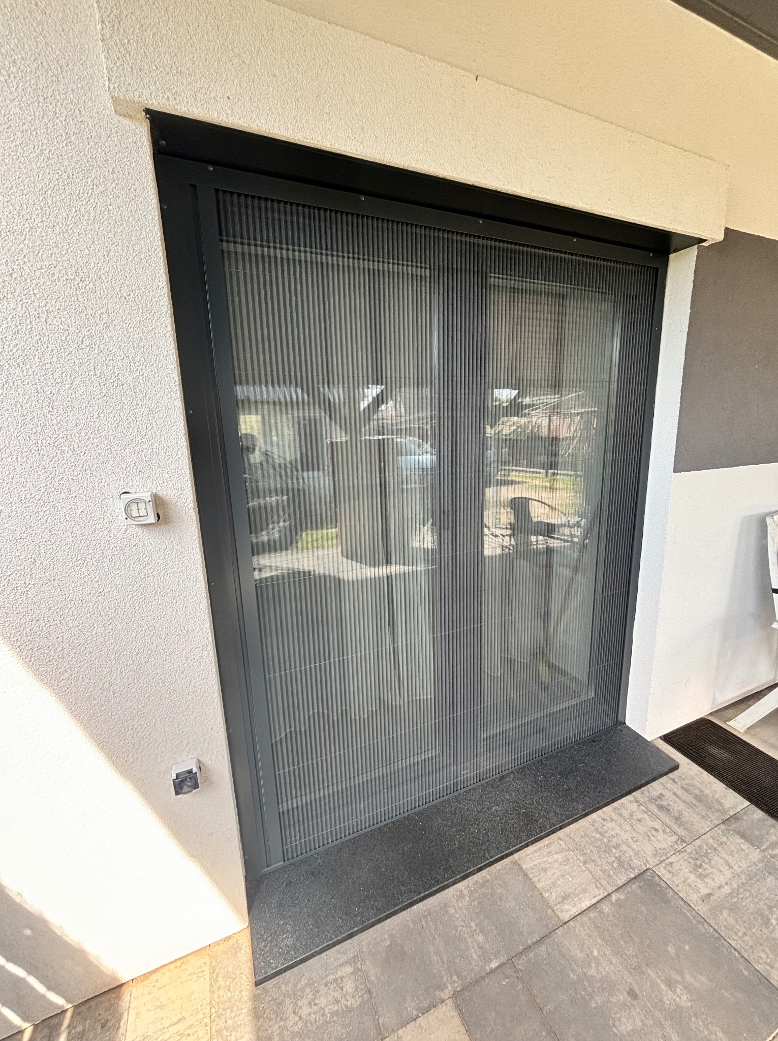 Zamknięta, plisowana moskitiera w czarnej ramie, zamontowana w drzwiach tarasowych, widok z zewnątrz, na szarej kostce brukowej.