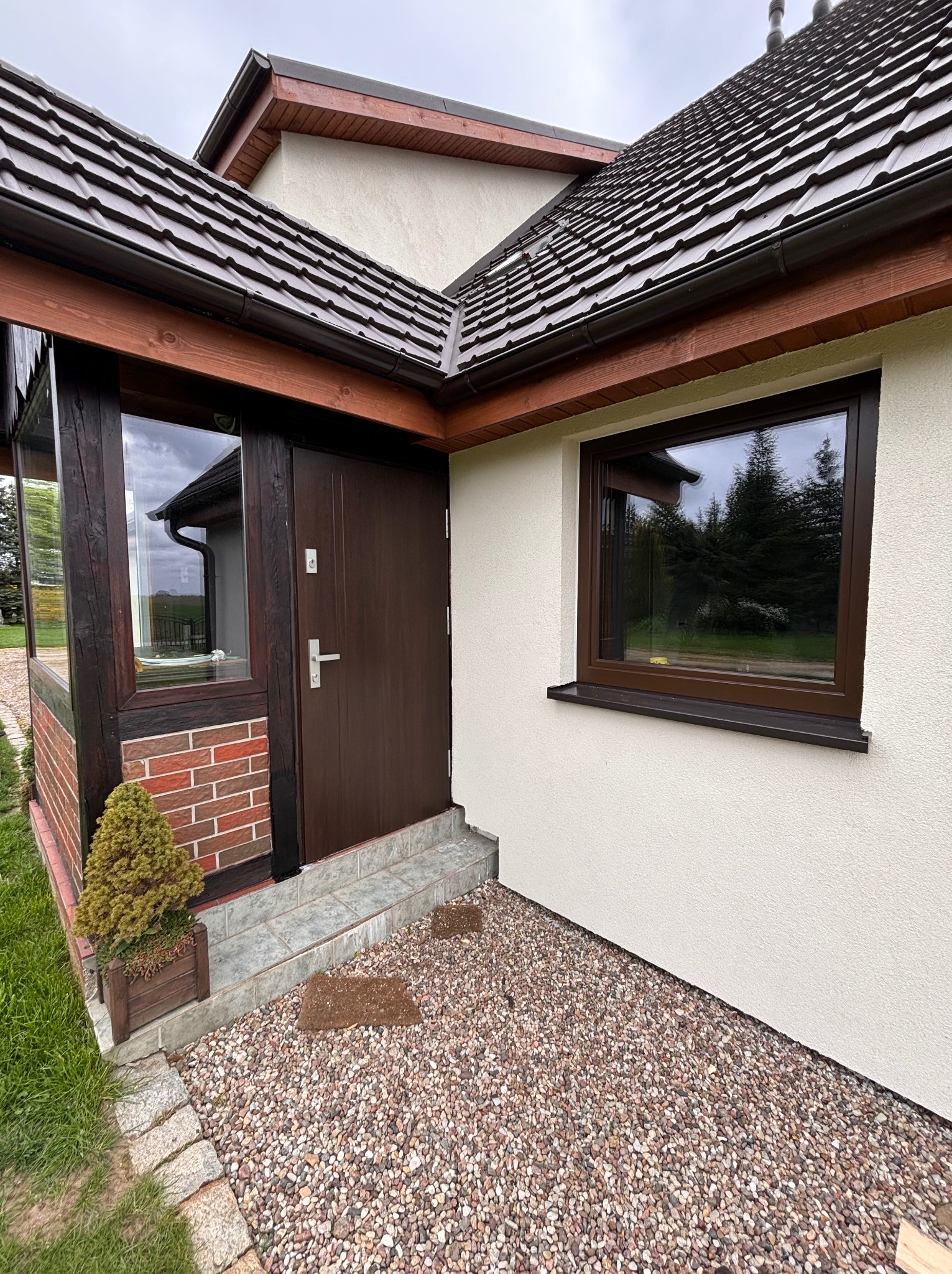 Brązowe okno PCV z parapetem z konglomeratu oraz ciemnobrązowe drzwi wejściowe w jasnym domu z kamiennym podłożem i małą rośliną w doniczce.