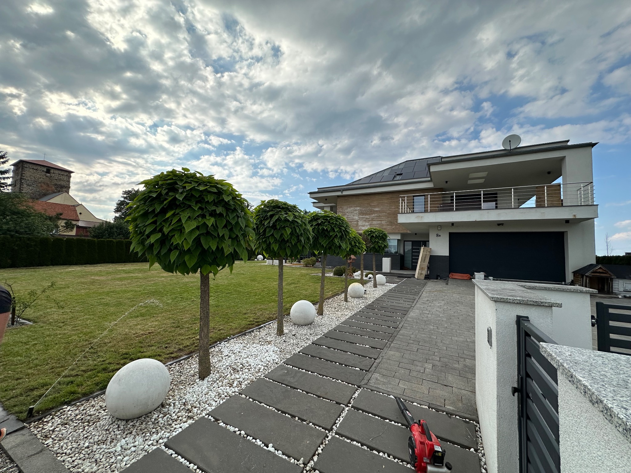 Nowoczesny ogród w Lesznie z równo przystrzyżonym trawnikiem, kulistymi drzewami otoczonymi białymi kamieniami, podlewaniem automatycznym i ścieżką z szarych płyt, prowadzącą do domu z panelami...