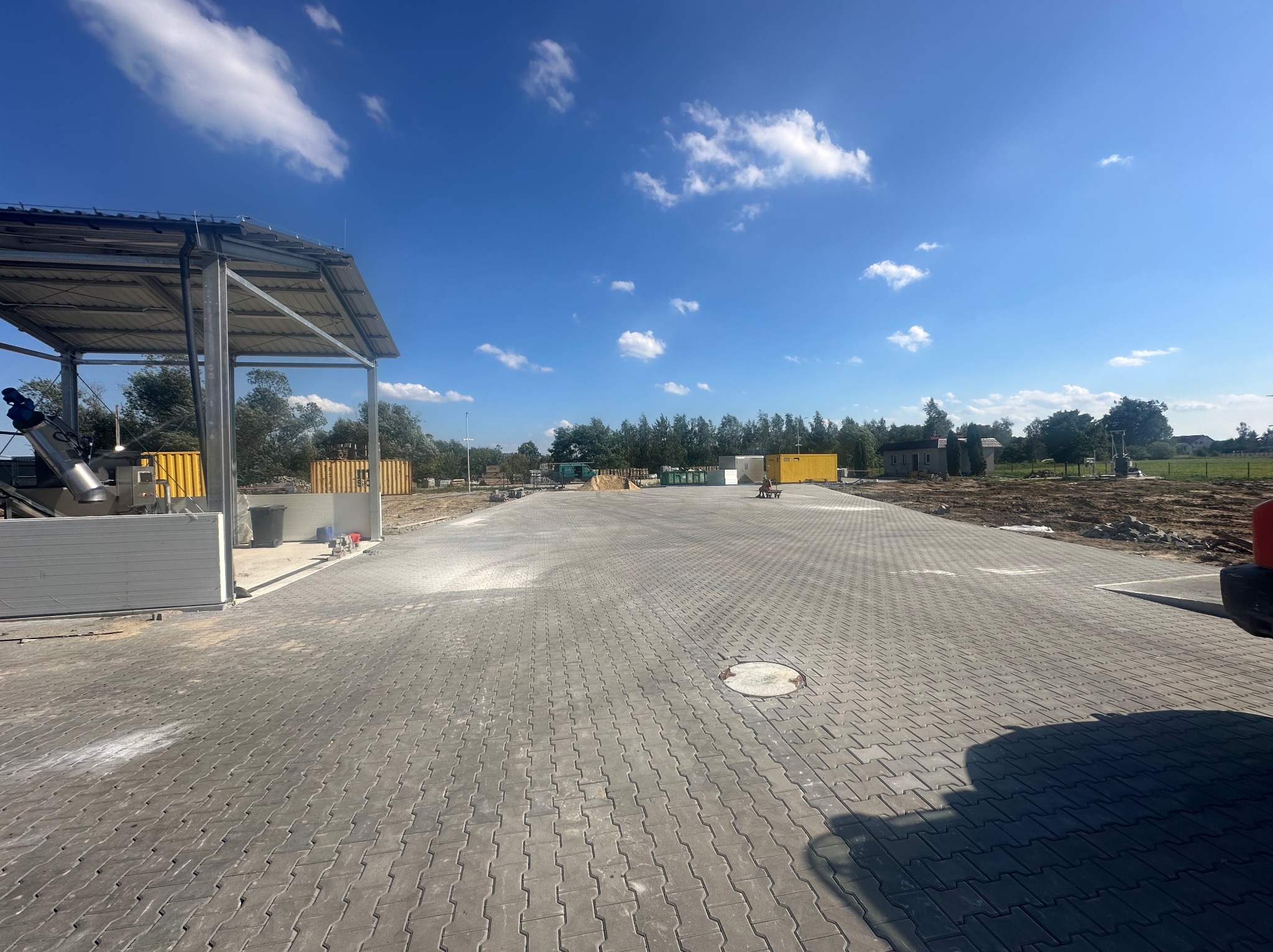 Rozległy plac wyłożony szarą kostką brukową, z widocznym fragmentem zadaszonej konstrukcji stalowej po lewej stronie i kontenerami w tle, pod błękitnym niebem z białymi obłokami.