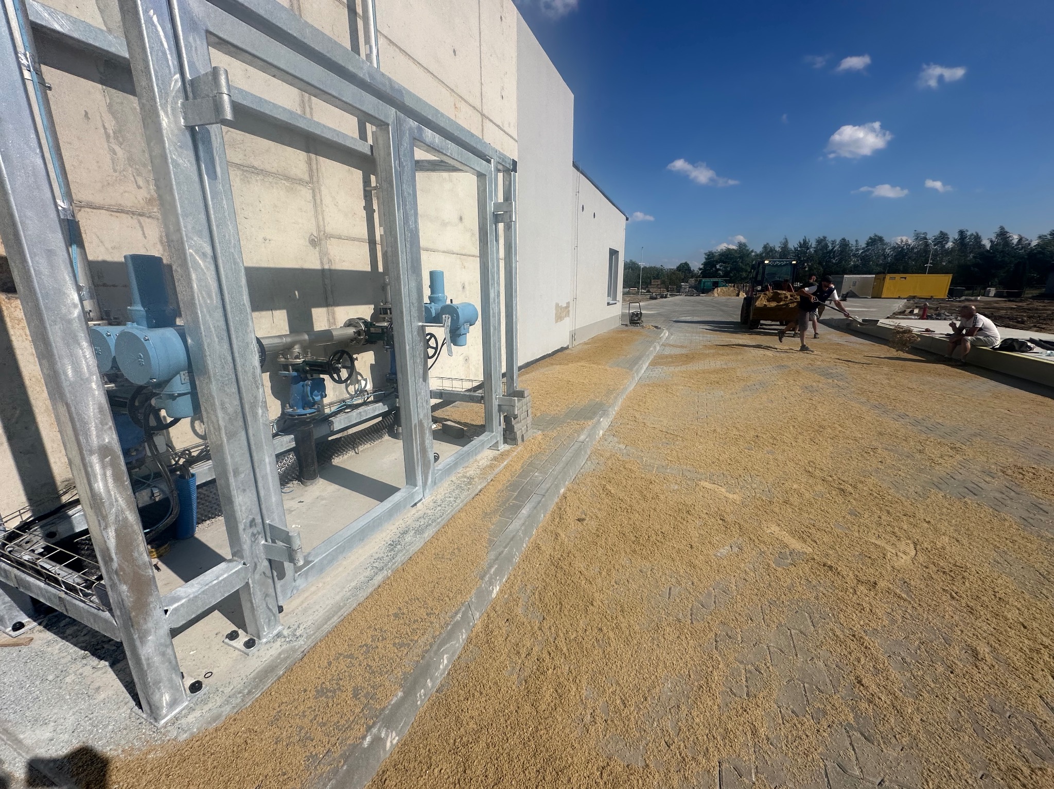 Przygotowanie podłoża pod kostkę brukową: rozsypywanie piasku na ułożonej kostce, widoczna stalowa konstrukcja, w tle pracownicy i maszyna budowlana.