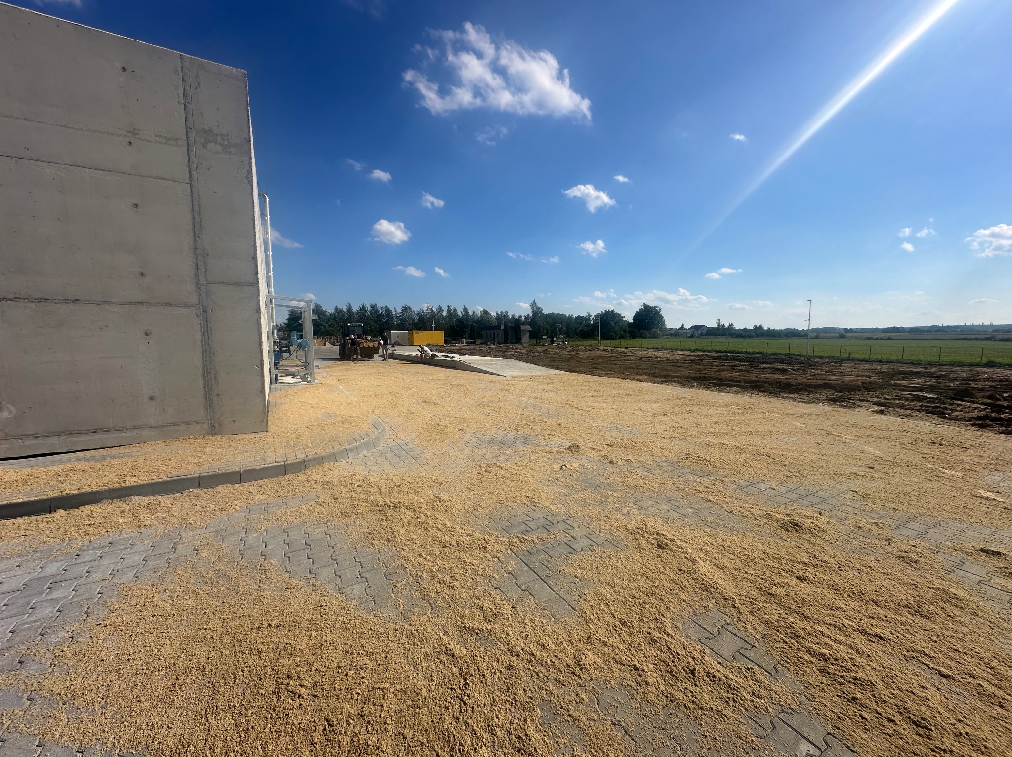 Nowo ułożona kostka brukowa pokryta warstwą piasku kwarcowego w celu wypełnienia szczelin, z widocznym fragmentem betonowego budynku i sprzętem budowlanym w tle, pod błękitnym niebem.