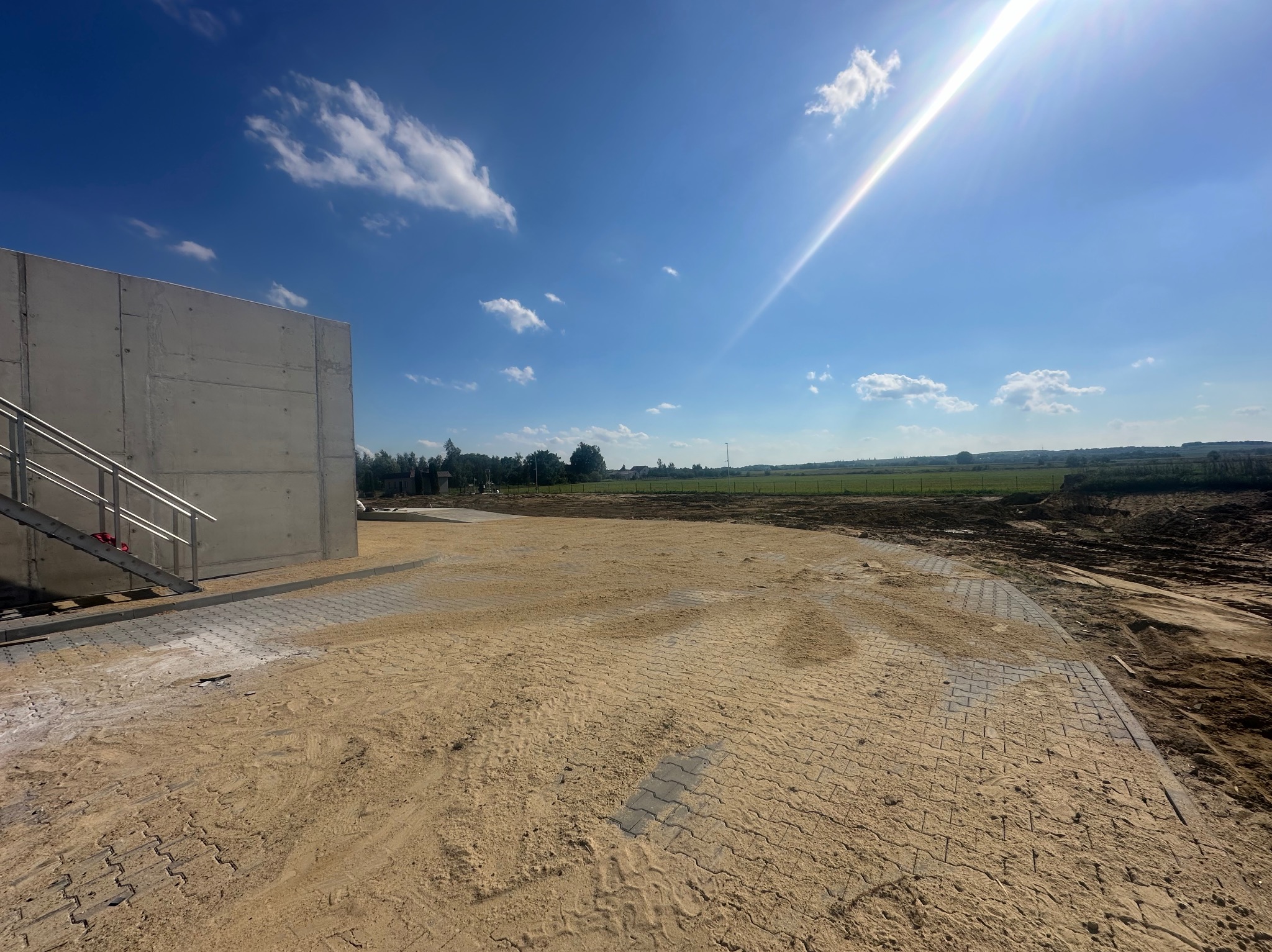 Układanie kostki brukowej w trakcie realizacji, widoczne fragmenty ułożonej kostki zasypane piaskiem, budynek z metalowymi schodami w tle, błękitne niebo z chmurami i słońcem.