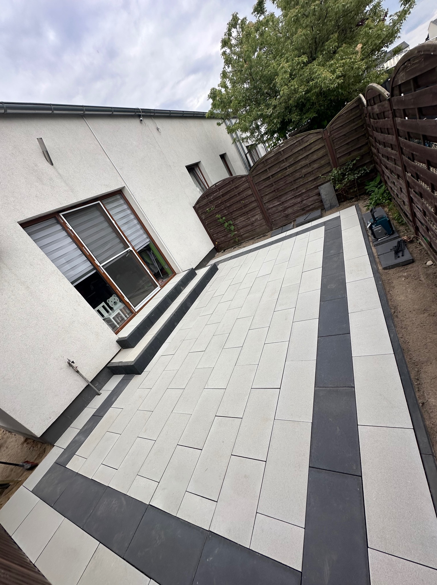 Nowo ułożony taras z jasnoszarej kostki brukowej z ciemnoszarą obwódką, prowadzący do schodów i okna domu, widoczny fragment drewnianego ogrodzenia i drzewa w tle.