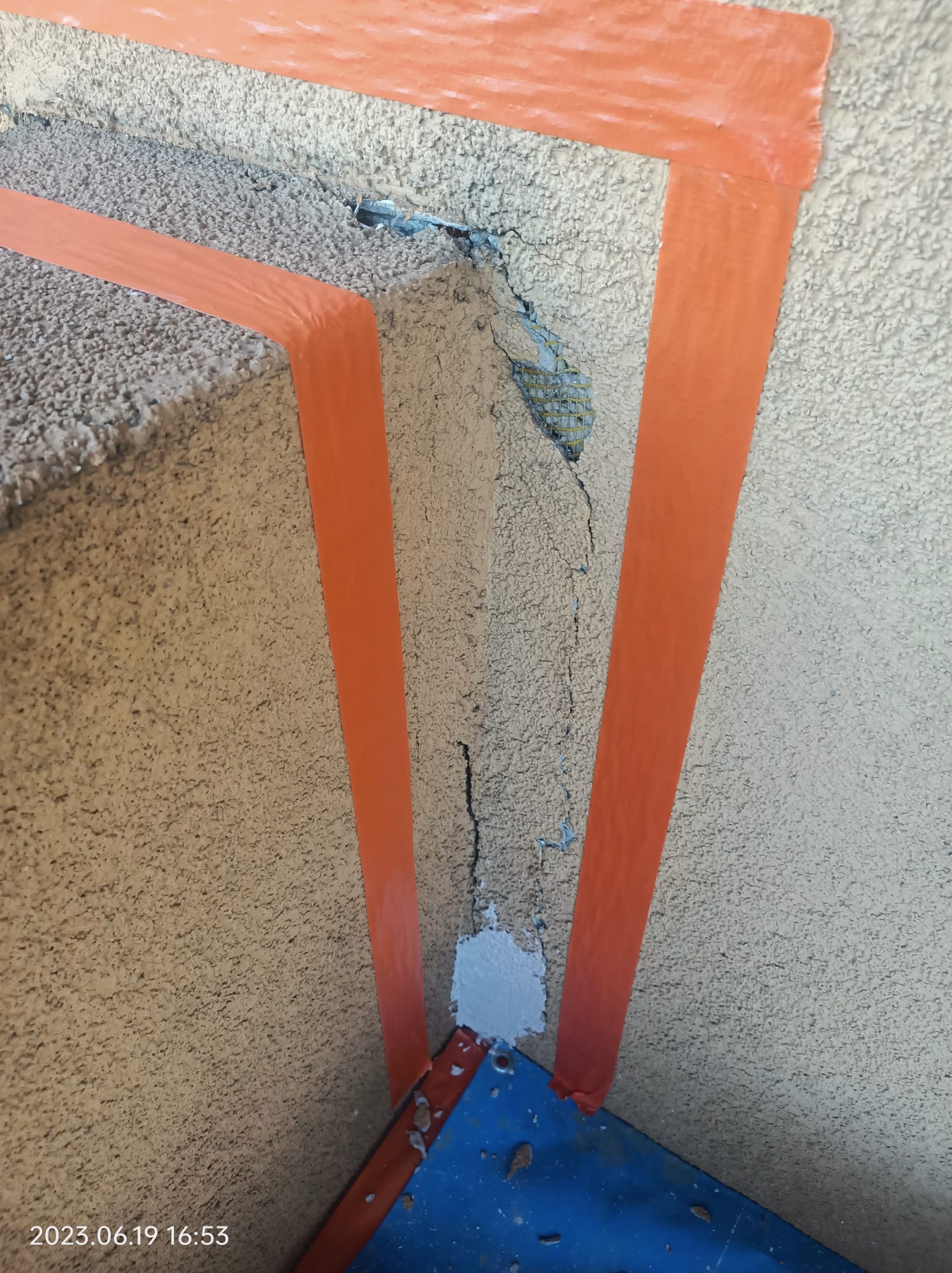 Przygotowanie do naprawy pękniętej elewacji z użyciem pomarańczowej taśmy malarskiej i niebieskiego narzędzia; widoczne uszkodzenia i odsłonięta siatka zbrojeniowa.