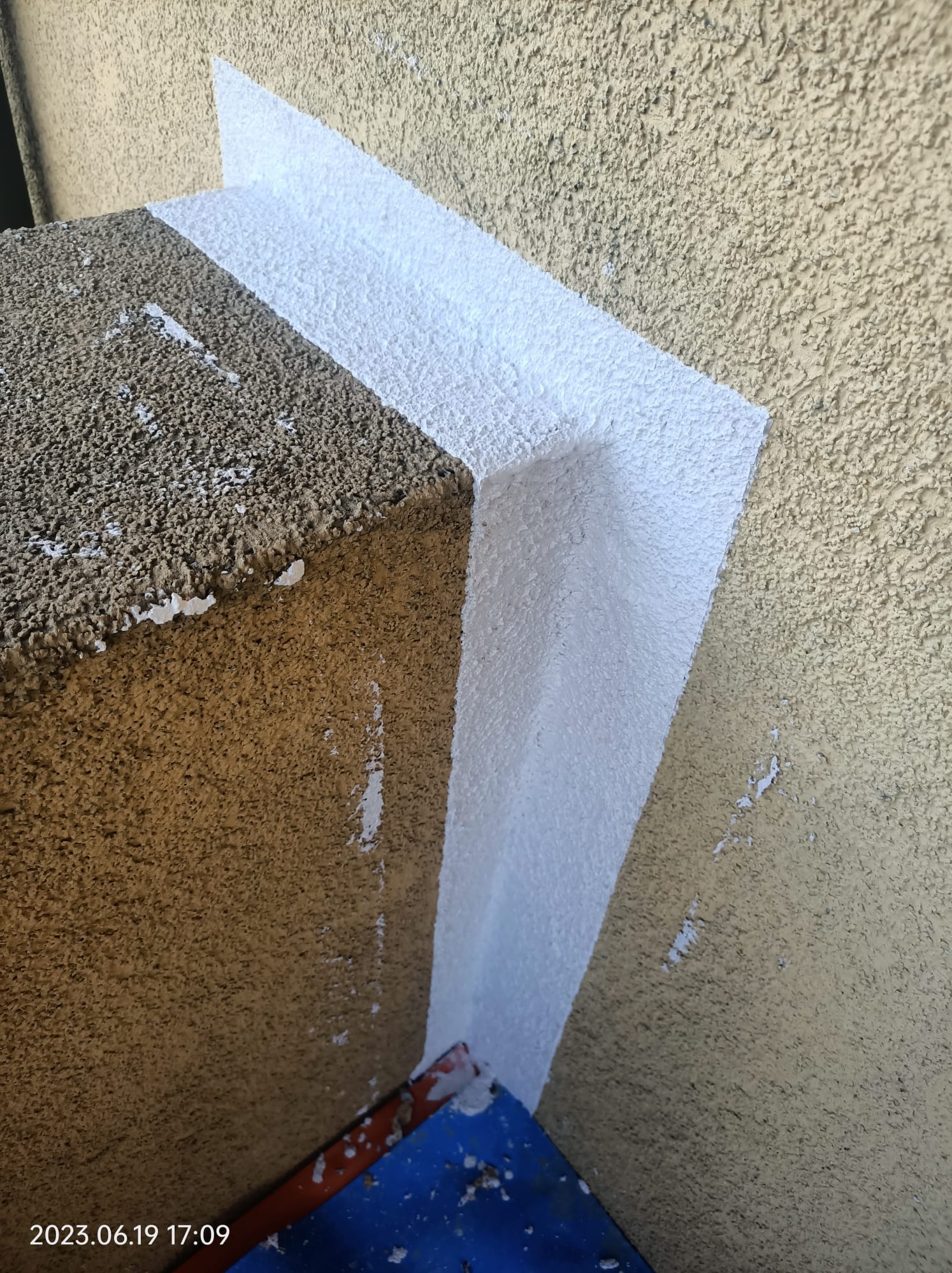 Fragment elewacji z widocznym świeżo pomalowanym, białym pasem przy krawędzi, z pozostałościami farby na sąsiedniej powierzchni i widocznym fragmentem kuwety malarskiej w rogu.