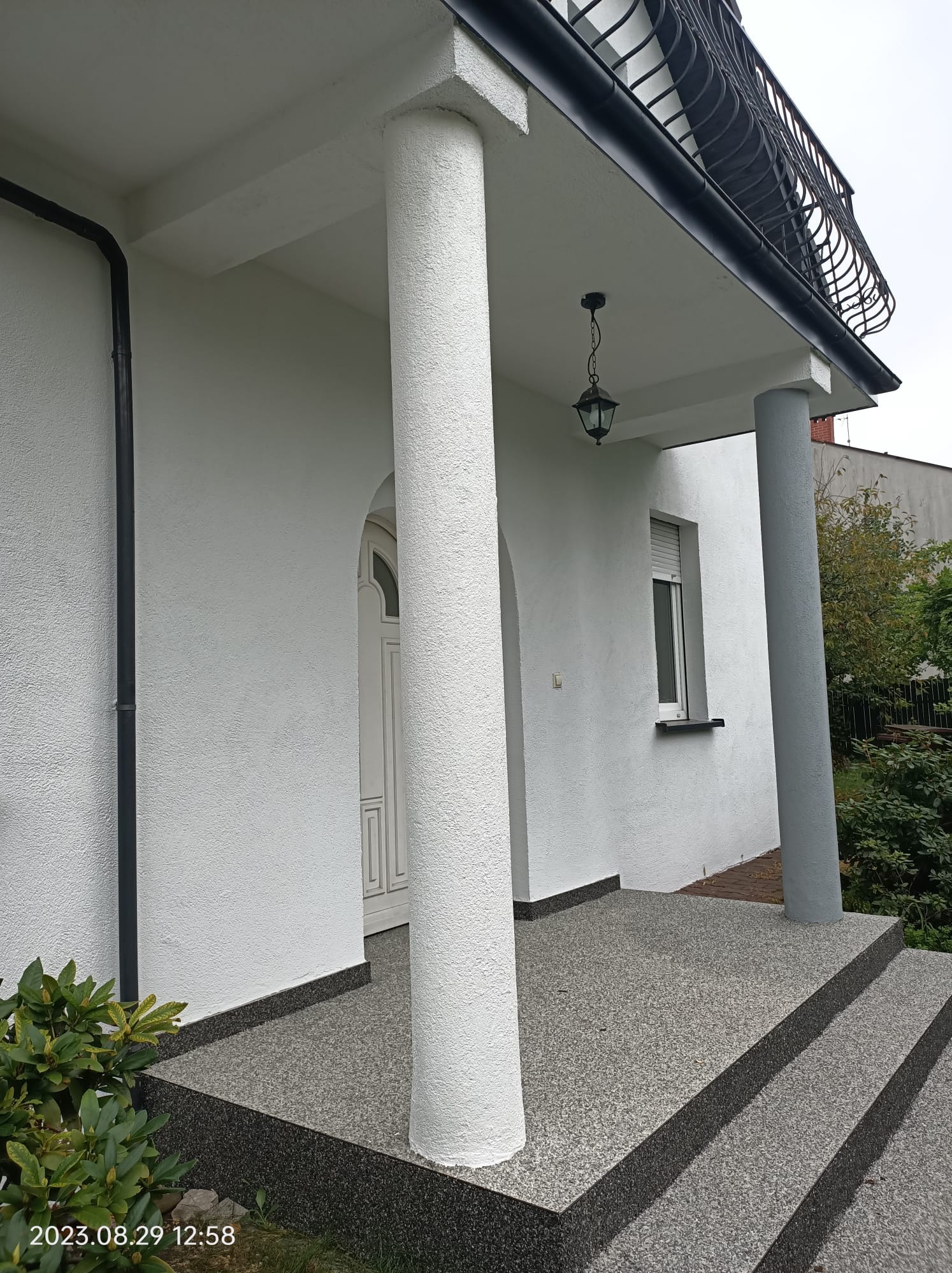Biała elewacja domu z kolumnami, widoczne schody wejściowe wykończone drobnym kamieniem, czarna rynna, lampa wisząca i balkon z ozdobną balustradą.
