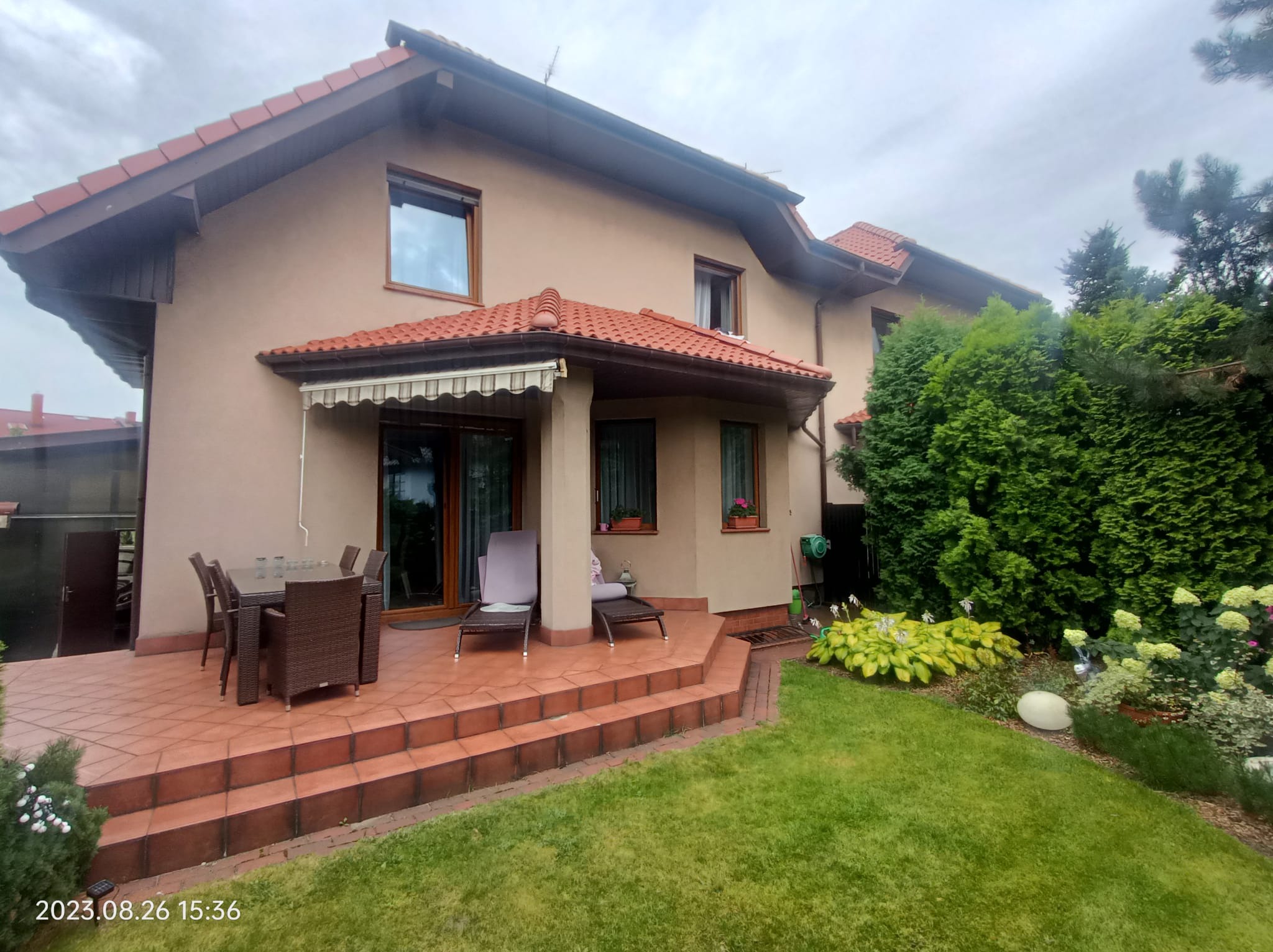 Dom z tarasem wyłożonym czerwoną terakotą, schodami i meblami ogrodowymi, z zadbanym trawnikiem i roślinnością.