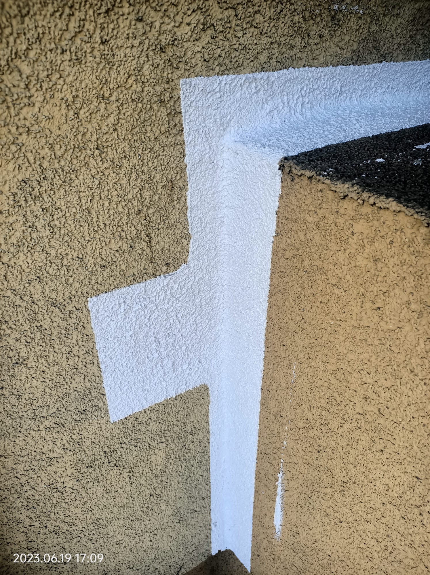 Fragment elewacji budynku z widocznym, świeżo nałożonym białym szpachlowaniem w narożniku, kontrastującym z chropowatą, beżową strukturą tynku.