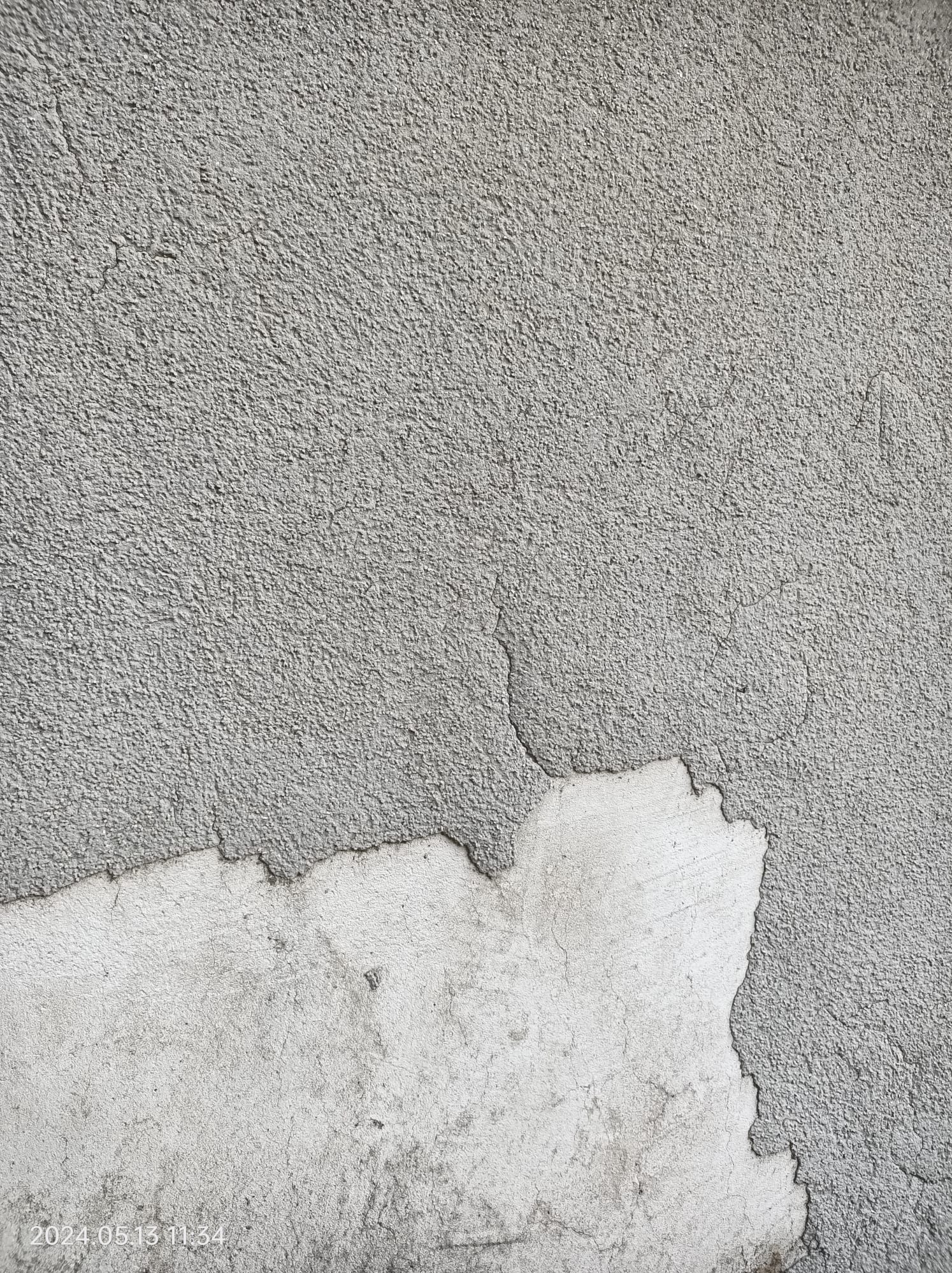 Fragment szarej, chropowatej elewacji z widocznym ubytkiem i odsłoniętą warstwą spodnią w odcieniu bieli. Widoczne pęknięcia na powierzchni.