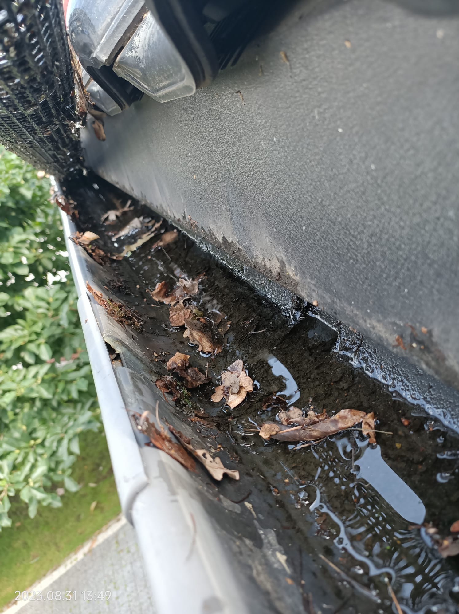 Zanieczyszczona rynna z liśćmi i stojącą wodą, widok z góry na tle zieleni.