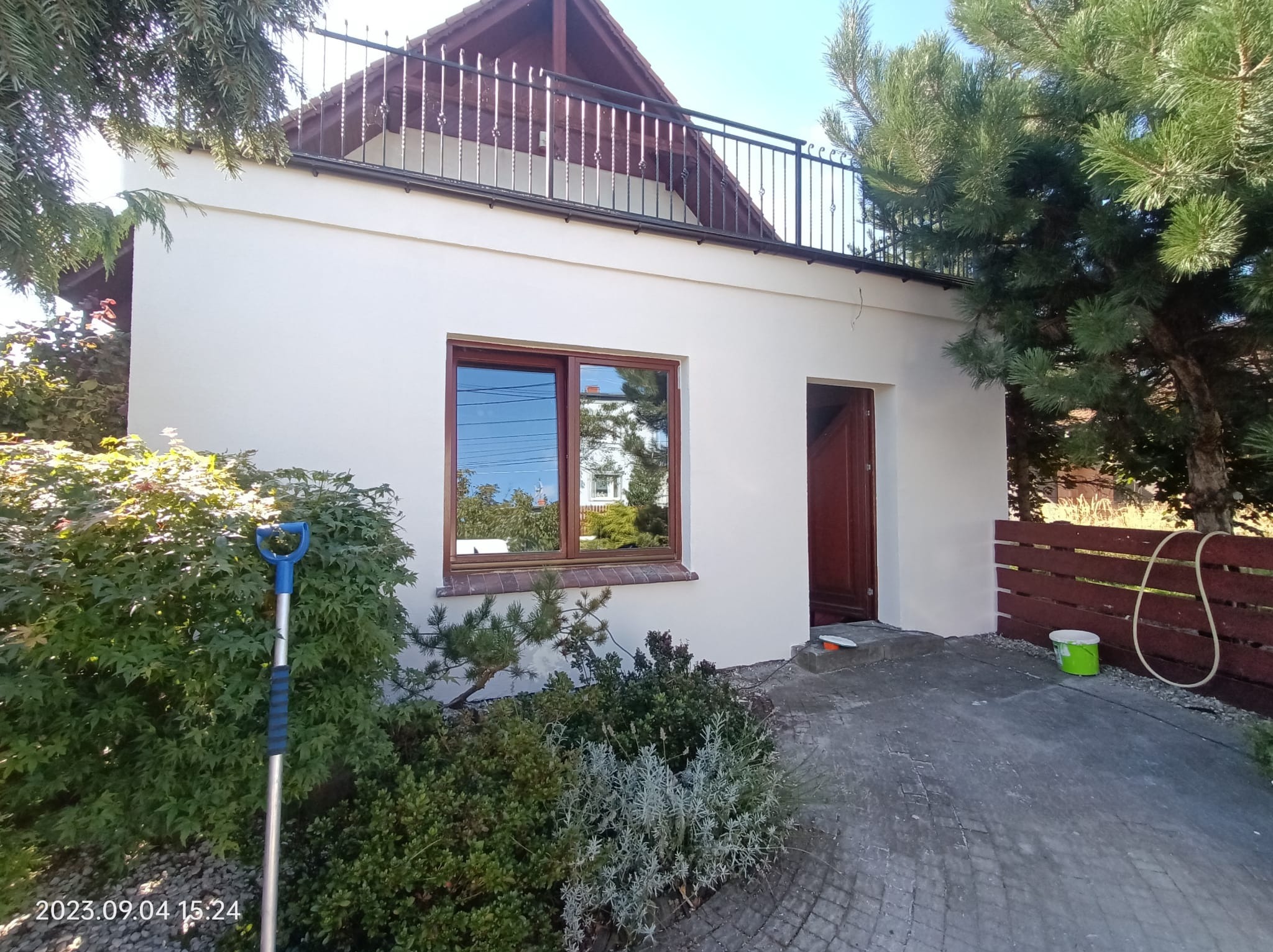 Biała elewacja budynku z brązowymi ramami okiennymi i drzwiami, widoczny fragment balkonu z czarną balustradą, zieleń w tle i na pierwszym planie, ślady świeżego malowania na chodniku.