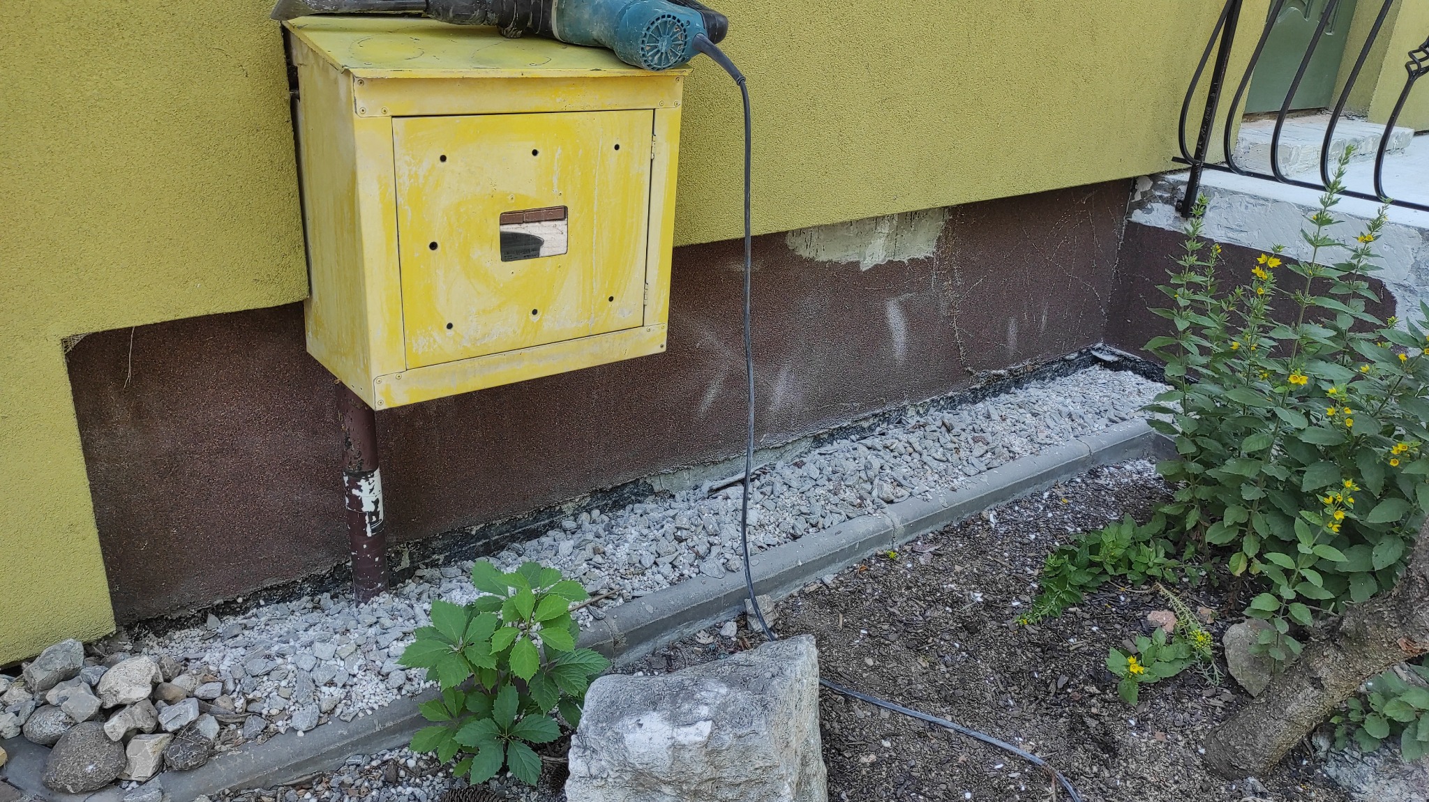 Przygotowanie do prac elewacyjnych: widoczny fragment ściany budynku z żółtą elewacją, brązową cokołem, skrzynka gazowa z leżącą na niej wiertarką oraz świeżo wysypany drenaż z kamyków przy...