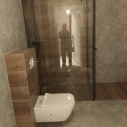 Onyx - Nowoczesna łazienka z kabiną prysznicową typu walk-in z czarnymi profilami, wisząca toaleta z zabudową, heksagonalne płytki na ścianach i podłodze, odbicie osoby w lustrze.