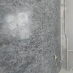 Szara ściana z efektem dekoracyjnym, widoczne gniazdko elektryczne z podłączonym kablem, rury przy ścianie.