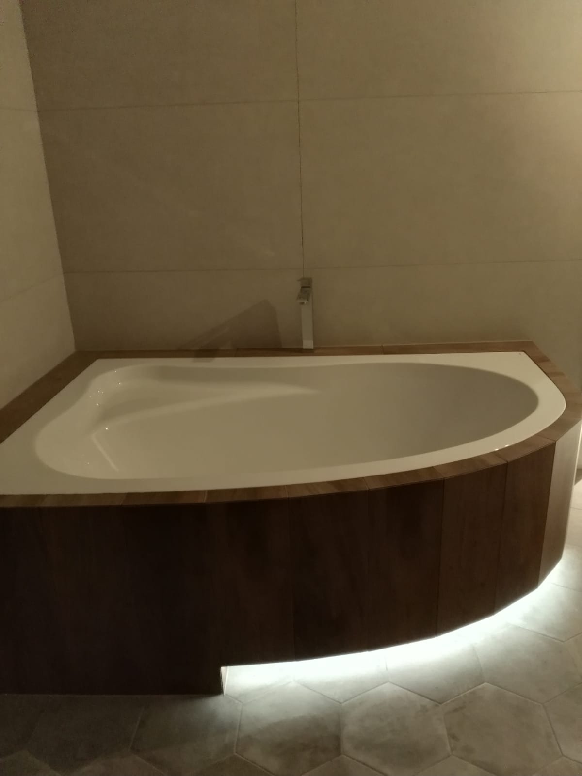 Wanna w obudowie z ciemnego drewna, podświetlona od dołu listwą LED, na podłodze szare heksagonalne płytki, bateria umywalkowa w tle.