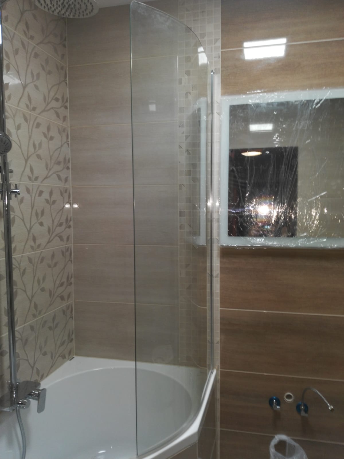 Zamontowana, narożna kabina prysznicowa z przezroczystego szkła, połączona z białą wanną. Na ścianach beżowe płytki z dekoracyjnym wzorem liści oraz płytki imitujące drewno. Lustro z folią ochronną.