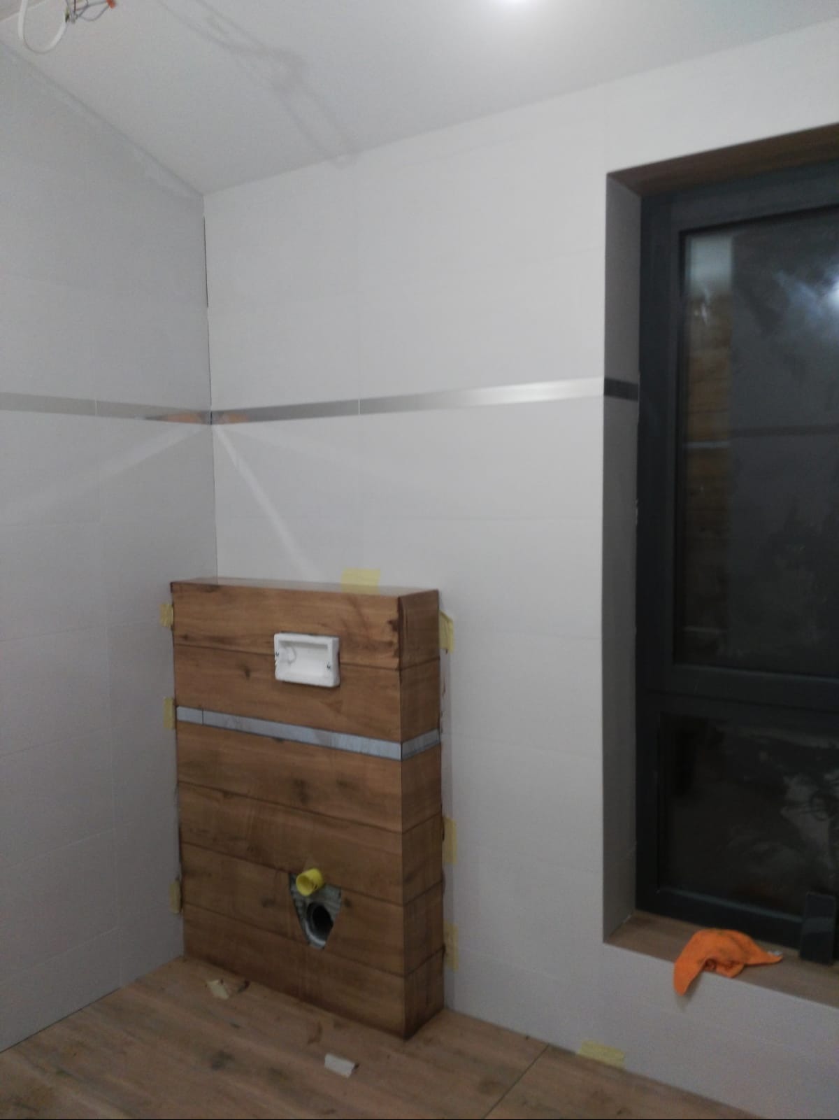 Drewniana obudowa stelaża podtynkowego WC w trakcie montażu, z widocznymi taśmami maskującymi na białych płytkach i drewnopodobnej podłodze, żółty element instalacji widoczny w otworze, okno...