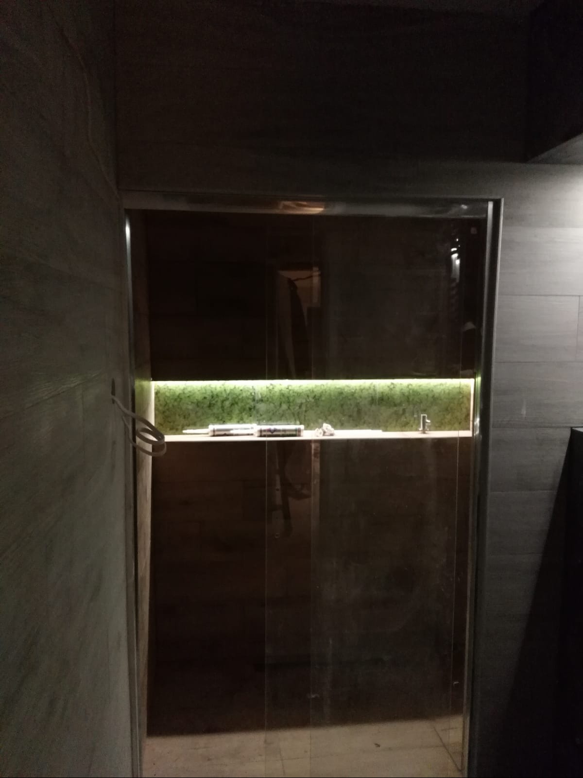 Szklana kabina prysznicowa z podświetlaną półką i widocznym aplikatorem silikonu, na tle szarych płytek imitujących kamień.