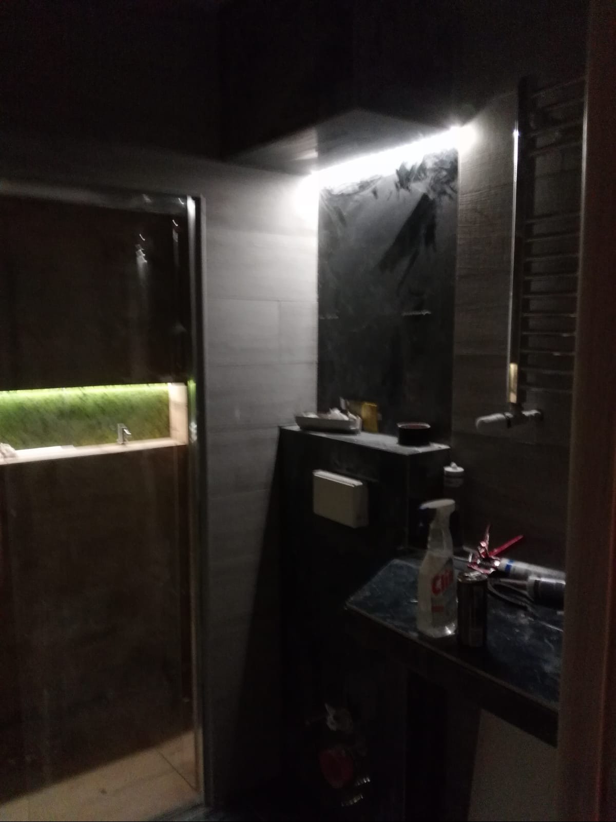 Ciemna łazienka w trakcie prac, widoczna kabina prysznicowa z podświetleniem LED, umywalka z narzędziami i środkami czystości, płytki na ścianach i grzejnik drabinkowy.