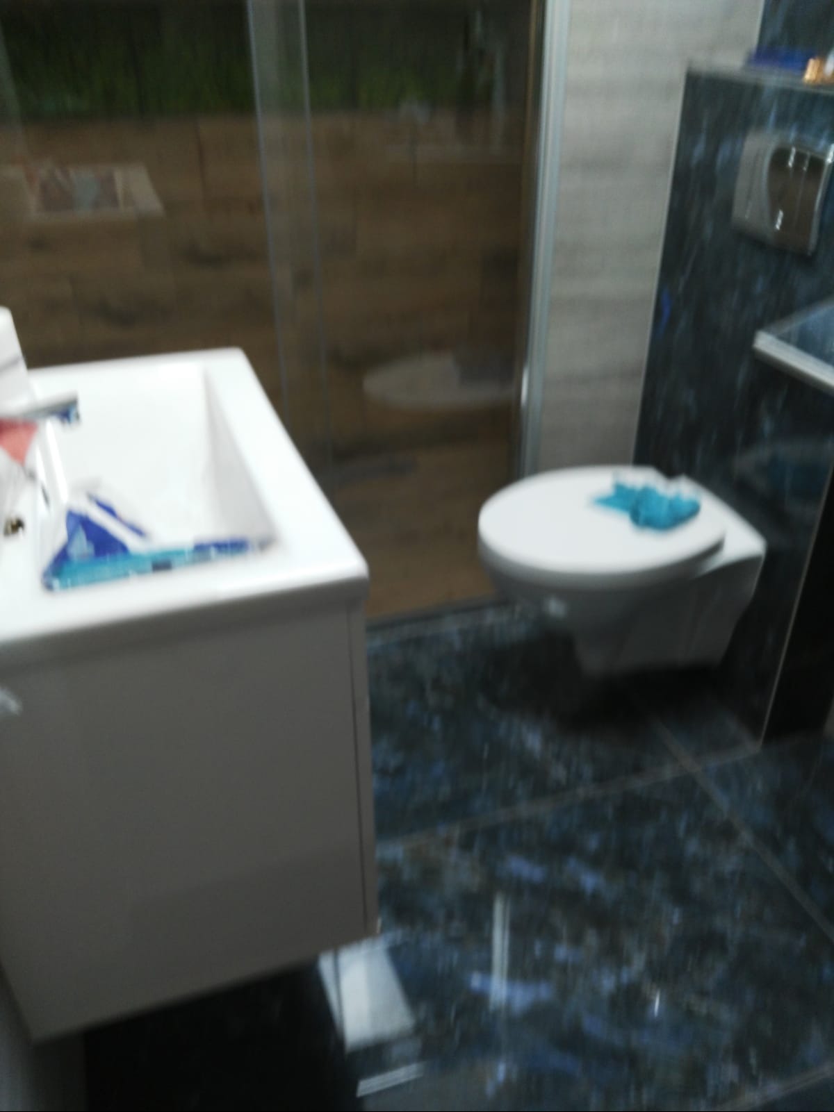 Umywalka z szafką, toaleta podwieszana z deską, kabina prysznicowa i płytki w odcieniach granatu i szarości tworzą nowoczesną aranżację łazienki.