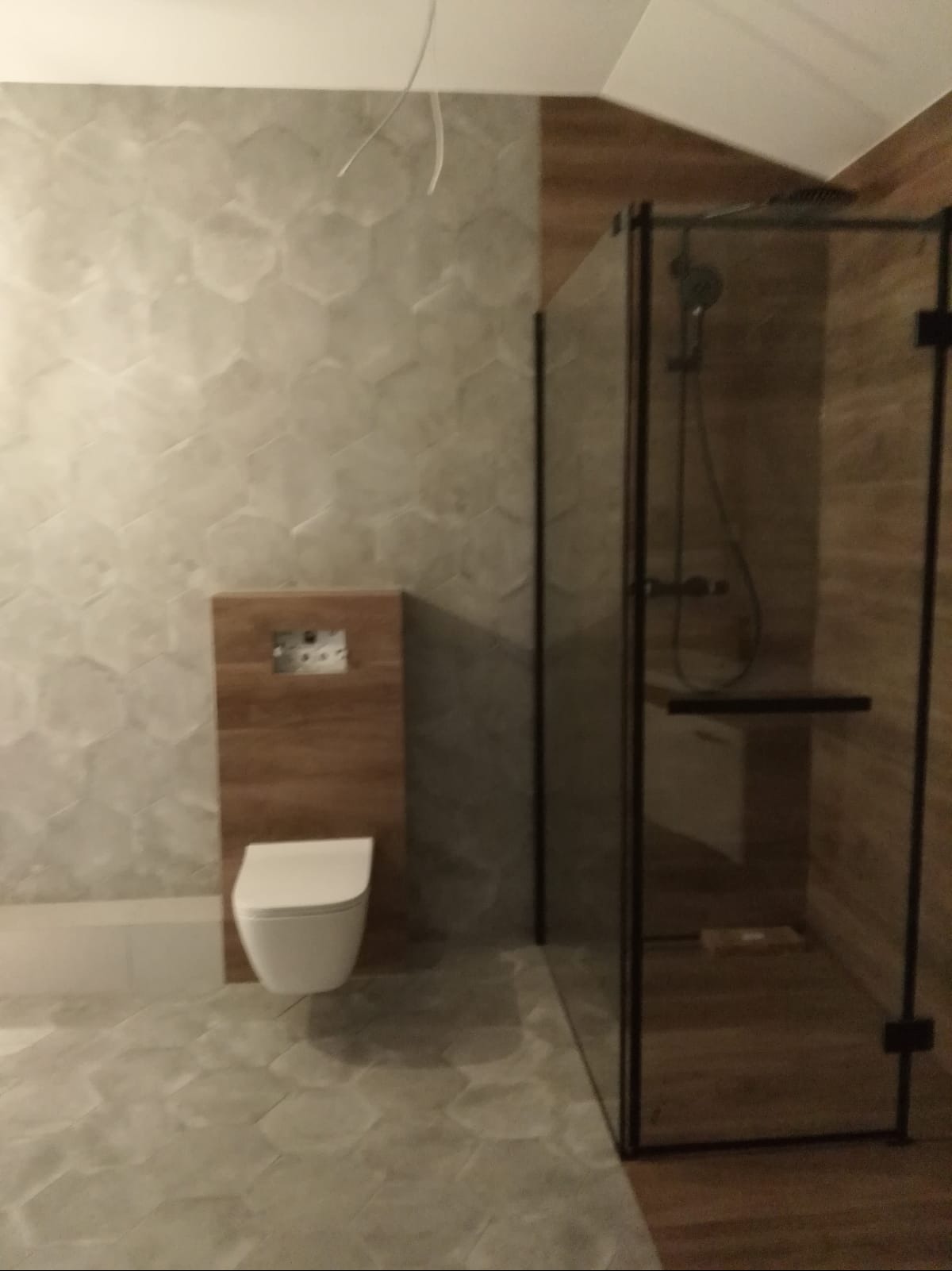 Nowoczesna łazienka z heksagonalnymi płytkami podłogowymi i ściennymi, zabudowana toaleta z drewnopodobną obudową oraz kabina prysznicowa z czarnymi profilami i drewnopodobnymi płytkami wewnątrz.