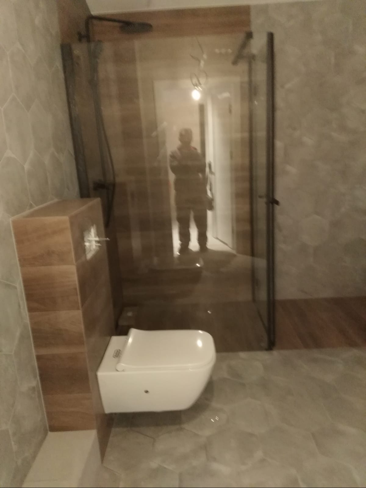 Nowoczesna łazienka z kabiną prysznicową typu walk-in z czarnymi profilami, wisząca toaleta z zabudową, heksagonalne płytki na ścianach i podłodze, odbicie osoby w lustrze.