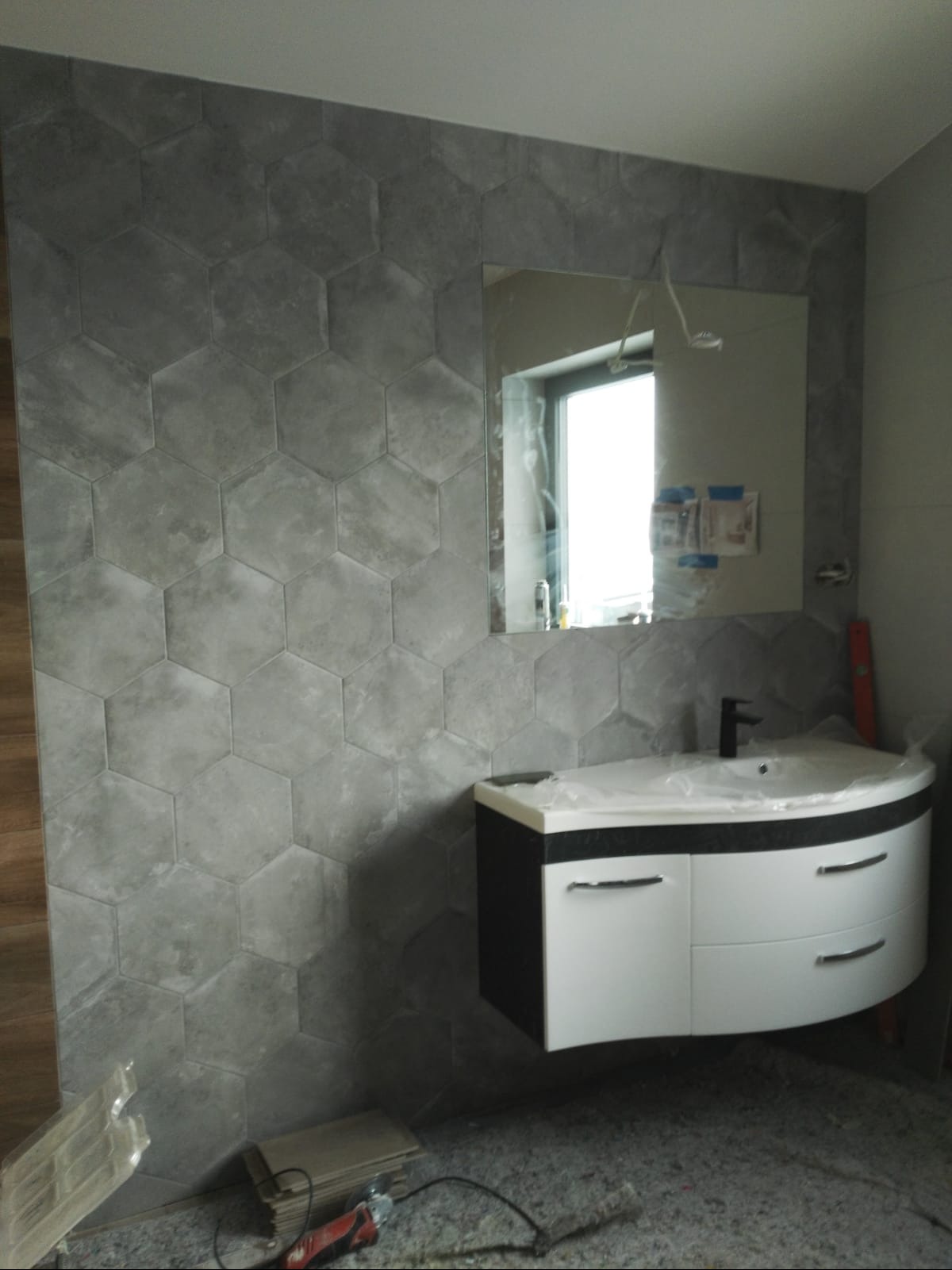 Szare heksagonalne płytki na ścianie łazienki, umywalka z szafką w kolorze czarno-białym, lustro, narzędzia na podłodze, poziomica przy ścianie.
