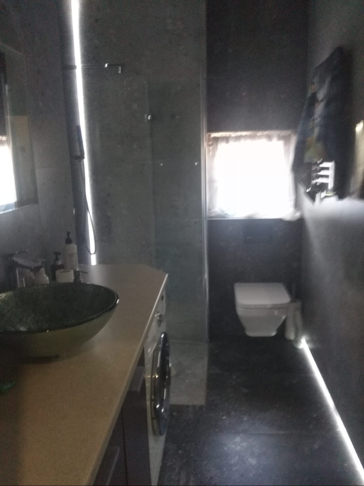 Nowoczesna łazienka z ciemnymi płytkami, szklaną kabiną prysznicową, umywalką na blacie o wzorzystym wnętrzu i pralką pod blatem.