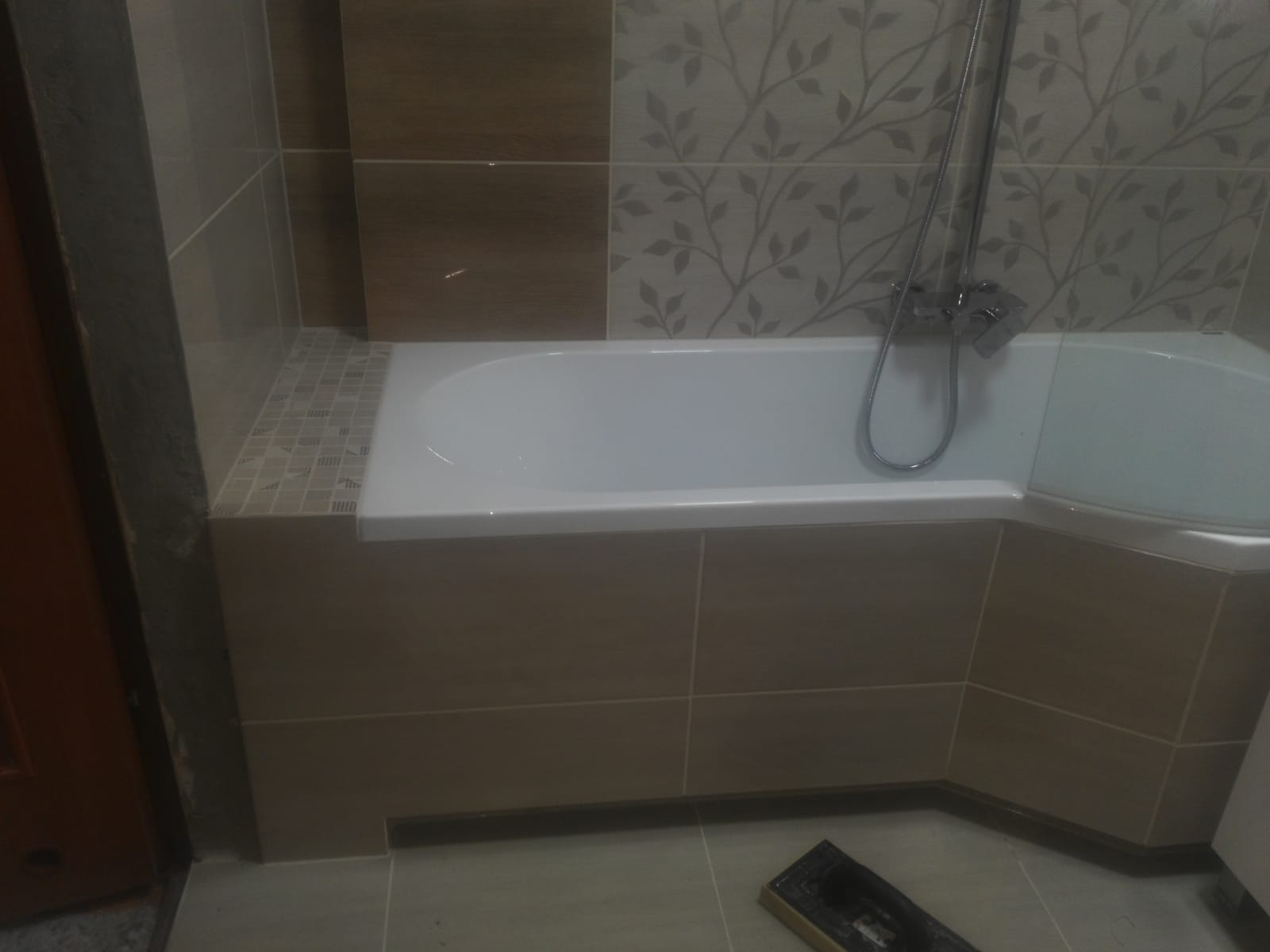 Biała wanna narożna z zamontowaną baterią, obudowana brązowymi płytkami, z dekoracyjnymi płytkami ściennymi z motywem roślinnym w tle.