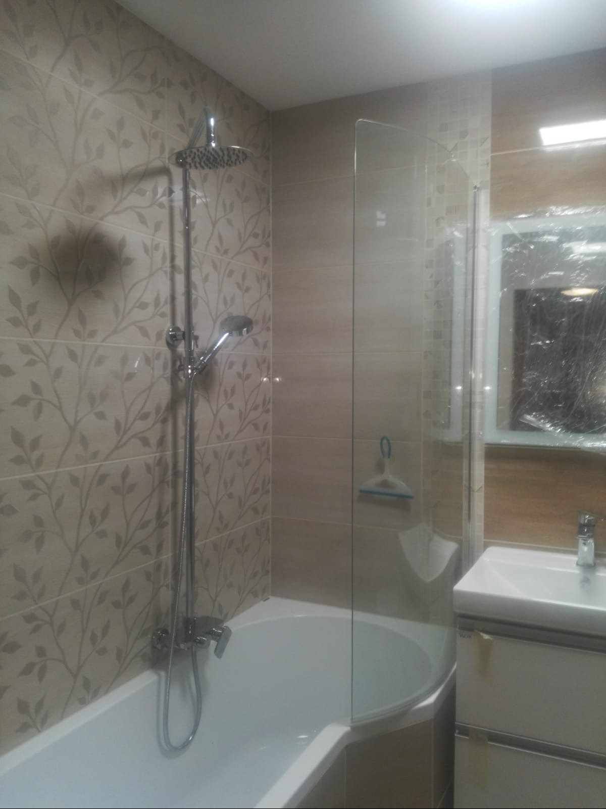 Wykończona łazienka z wanną, szklanym parawanem nawannowym, umywalką z szafką i baterią. Ściany wyłożone płytkami w dwóch odcieniach beżu, jedna ze wzorem roślinnym.