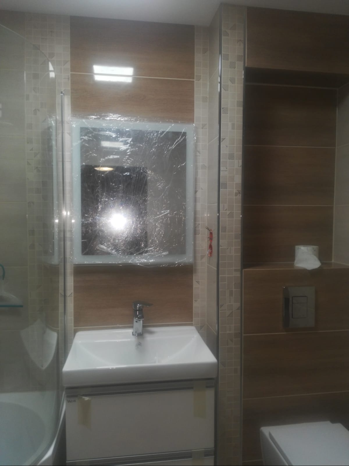 Nowoczesna łazienka z umywalką i szafką w zabudowie, lustro zabezpieczone folią ochronną, płytki imitujące drewno na ścianach, widoczna toaleta i kabina prysznicowa.