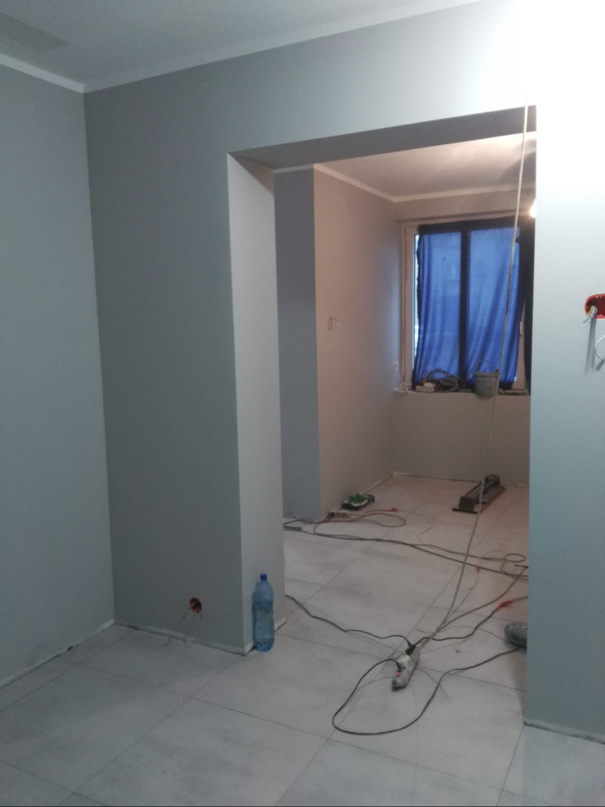 Szare ściany w pokoju po malowaniu, widoczne kable i narzędzia na podłodze, okno zasłonięte niebieską tkaniną, butelka wody przy ścianie.