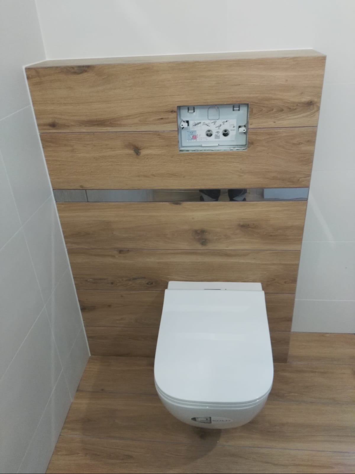 Podwieszana toaleta z zabudową w drewnopodobnej okładzinie z metalowym akcentem, widoczny stelaż podtynkowy przed montażem przycisku spłukującego, białe płytki na ścianie po lewej stronie, podłoga...