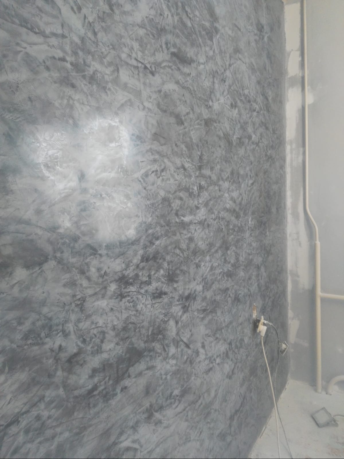 Szara ściana z efektem dekoracyjnym, widoczne gniazdko elektryczne z podłączonym kablem, rury przy ścianie.