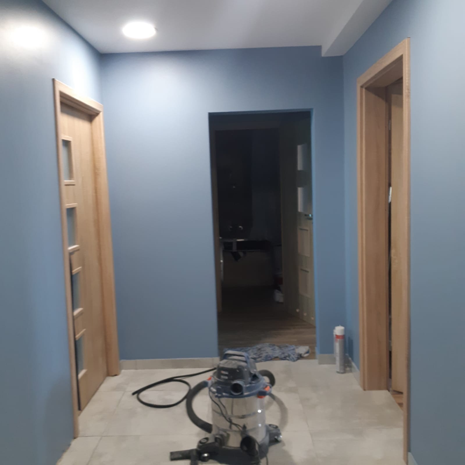 Korytarz po malowaniu na niebiesko, z zamontowanymi drzwiami z drewnianą ościeżnicą, widoczny odkurzacz przemysłowy i resztki po pracy.