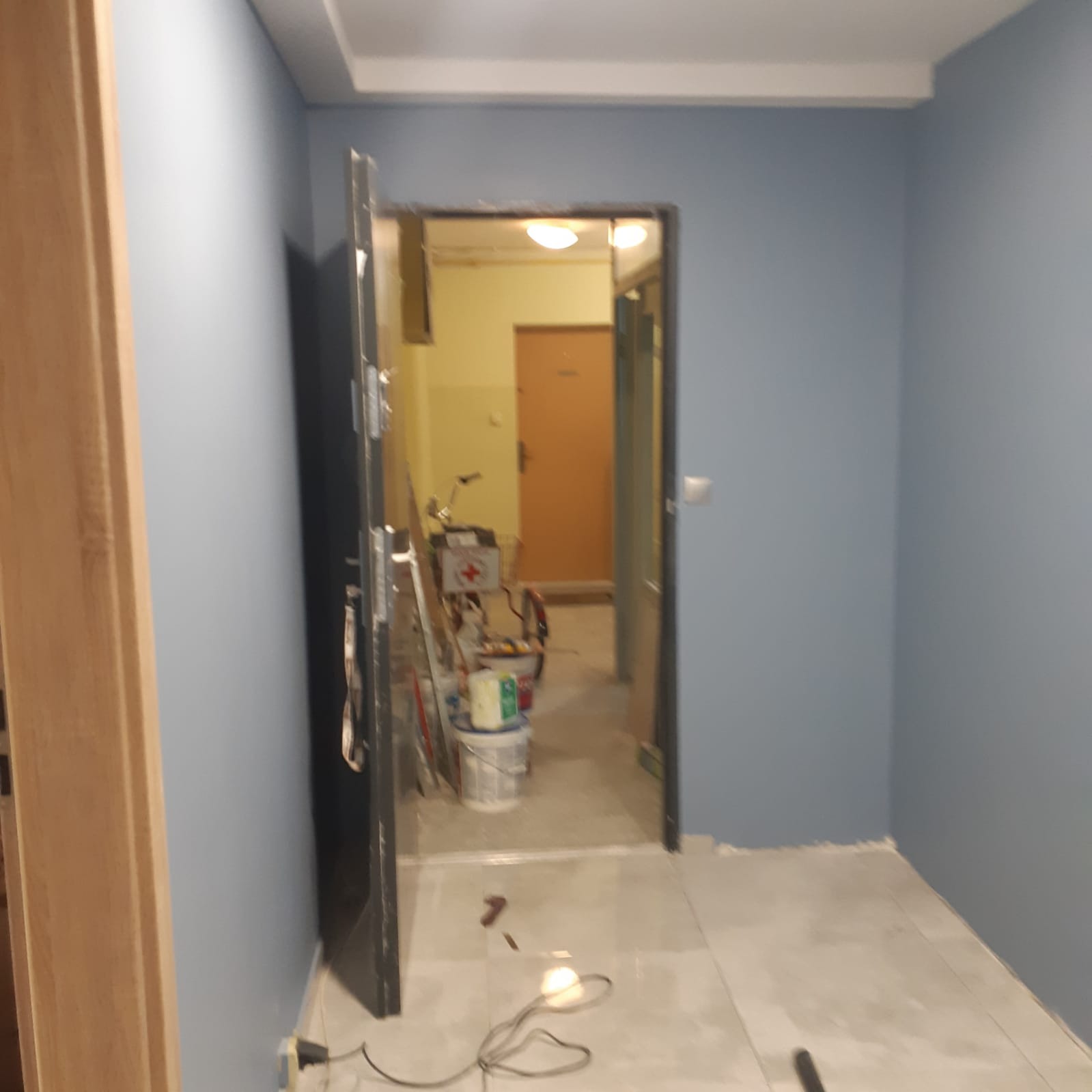 Świeżo pomalowane na niebiesko ściany widoczne przez otwarte drzwi. W tle widoczne narzędzia malarskie i rower w pokoju. Na podłodze kable.