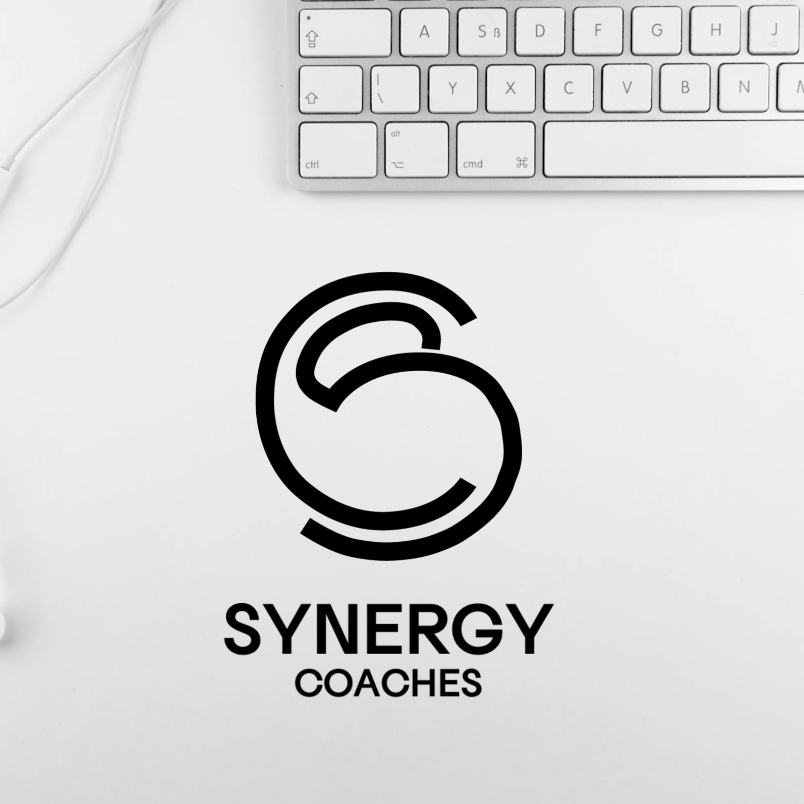 Minimalistyczne logo Synergy Coaches na białym biurku obok klawiatury Apple i białych słuchawek dousznych.