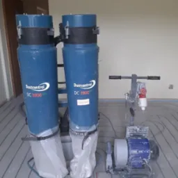 Dwa niebieskie odkurzacze przemysłowe Dustcontrol DC 3900 eco z zamocowanymi workami na wózku, stojące obok szlifierki do betonu na tle instalacji rurowej w podłodze.