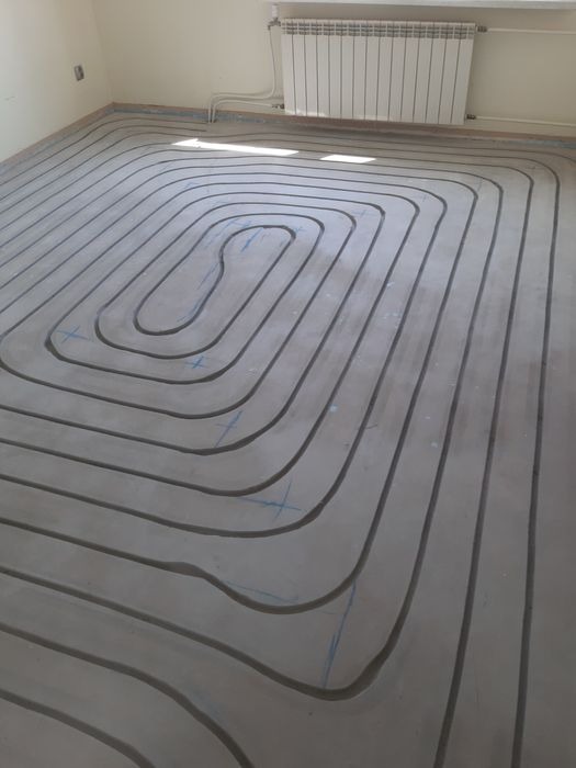 Wykonywanie instalacji grzewczej w podłodze, widoczne rury ułożone w pętle przed zalaniem wylewką, niebieskie oznaczenia na podłożu, grzejnik na ścianie w tle.