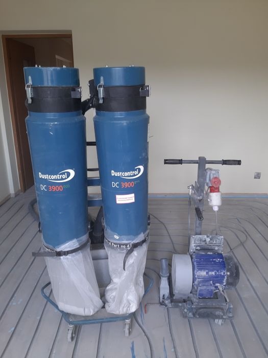 Dwa niebieskie odkurzacze przemysłowe Dustcontrol DC 3900 eco z zamocowanymi workami na wózku, stojące obok szlifierki do betonu na tle instalacji rurowej w podłodze.