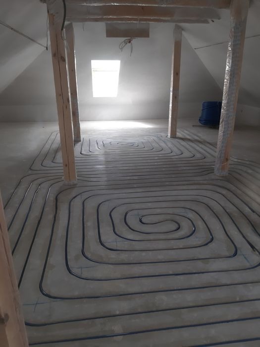 Rury ogrzewania podłogowego ułożone spiralnie w surowym pomieszczeniu na poddaszu, widoczne drewniane słupy zabezpieczone folią i okno dachowe.